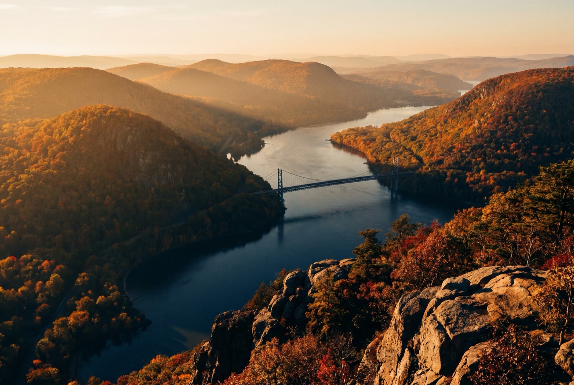 The Hudson Highlands at golden hour, the Hudson River winding between autumn mountains, seen from a rocky overlook