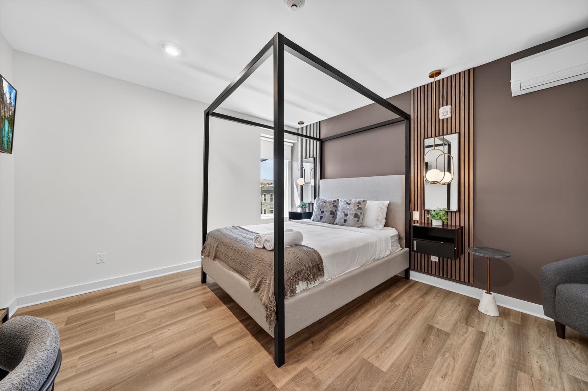 A standard suite at 52 Cottage Street: a black four-poster bed with white linen and a gray throw, a mauve feature wall with a wood-slat panel and wall sconces, a window on the left, and light wood floors